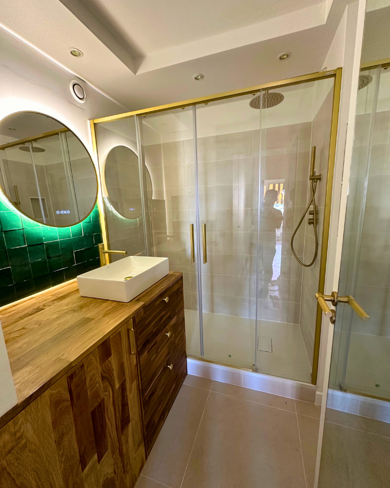Salle de bain moderne avec meuble-lavabo en bois, miroir rond, vasque et cabine de douche en verre avec des accents dorés. Des carreaux verts et un éclairage encastré sont visibles.