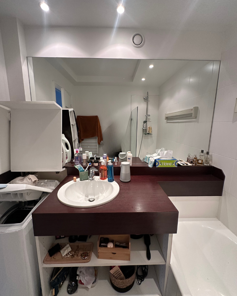 Salle de bain avec un grand miroir, un lavabo et un comptoir sombre. Des articles de toilette et des outils de toilettage sont éparpillés sur le comptoir. Une baignoire et une douche sont visibles en arrière-plan.