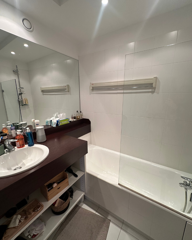 Une petite salle de bain avec un lavabo, un miroir et un ensemble douche-baignoire. Les produits de toilette sont sur des étagères et autour du lavabo. Des carreaux blancs recouvrent les murs.