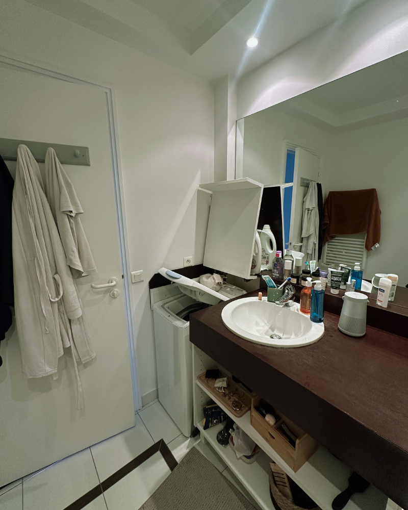Salle de bain moderne avec meuble-lavabo en bois, miroir rond, vasque et cabine de douche en verre avec des accents dorés. Des carreaux verts et un éclairage encastré sont visibles.
