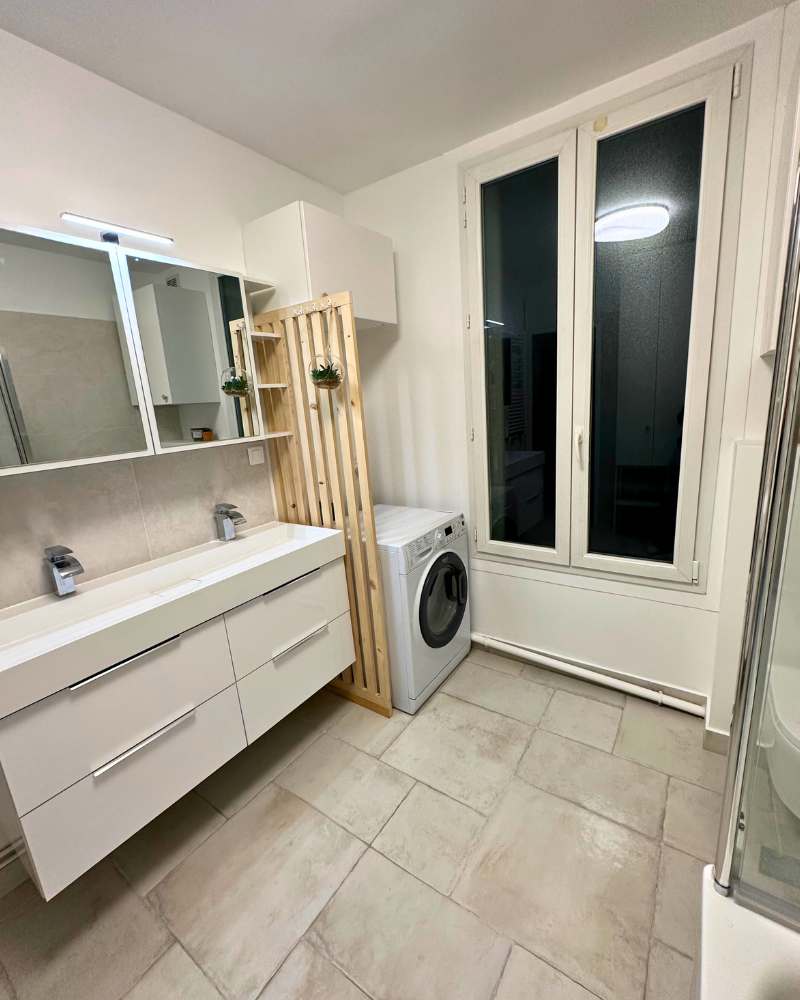 Salle de bain moderne avec double vasque, lave-linge, étagères en bois, grande fenêtre et sol carrelé.