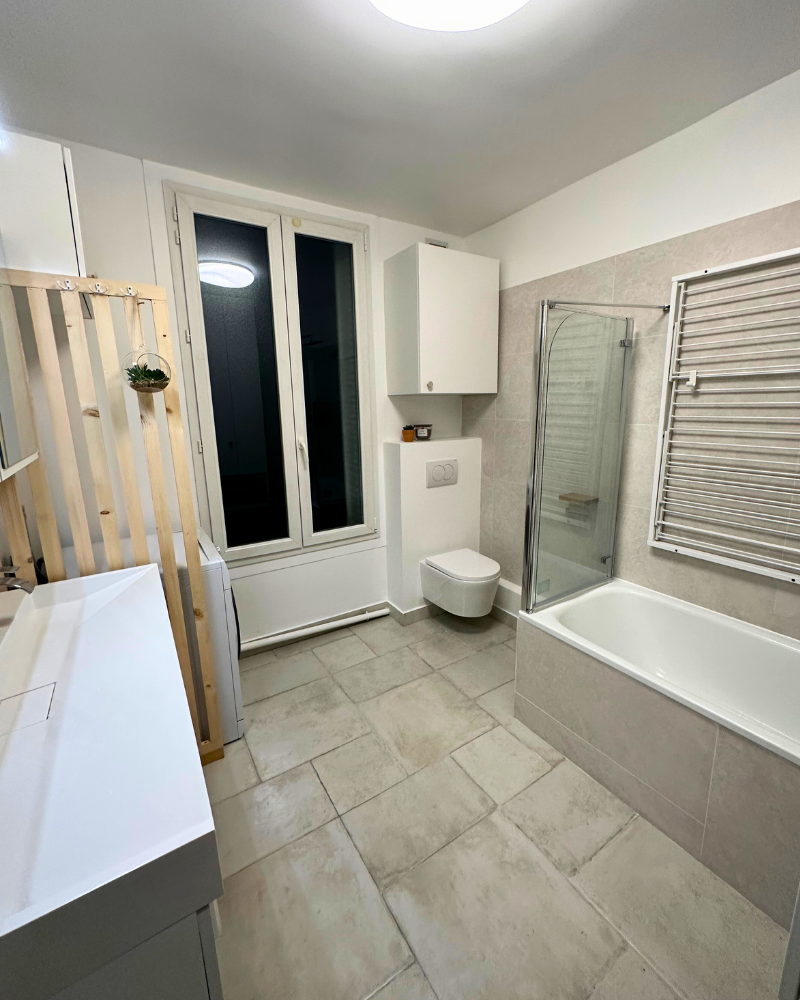 Une salle de bain moderne avec sol carrelé, baignoire avec pare-baignoire en verre, WC suspendu, lavabo et fenêtre. On peut y voir un sèche-serviettes et une petite plante suspendue à un séparateur en bois.