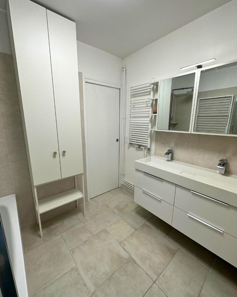 Salle de bain moderne avec armoires blanches, lavabo mural, armoire à glace, chauffe-serviettes et sol carrelé beige.