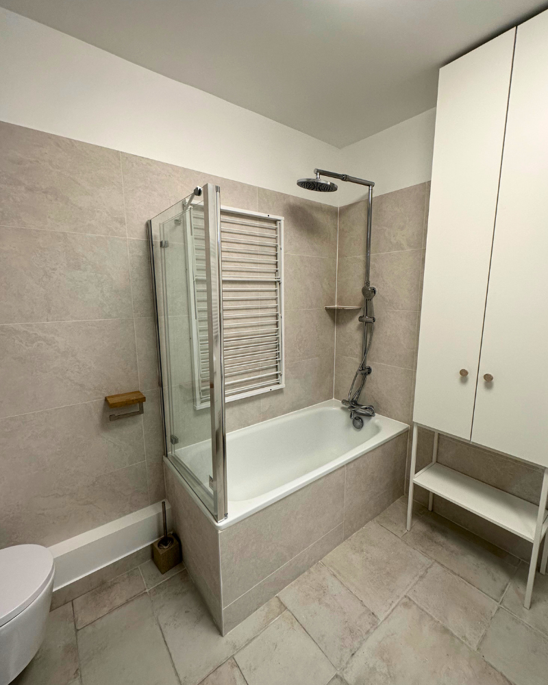 Salle de bain moderne avec baignoire-douche, paroi vitrée, radiateur mural et meuble de rangement blanc. Les murs et le sol carrelés beige clair créent un look épuré et spacieux.