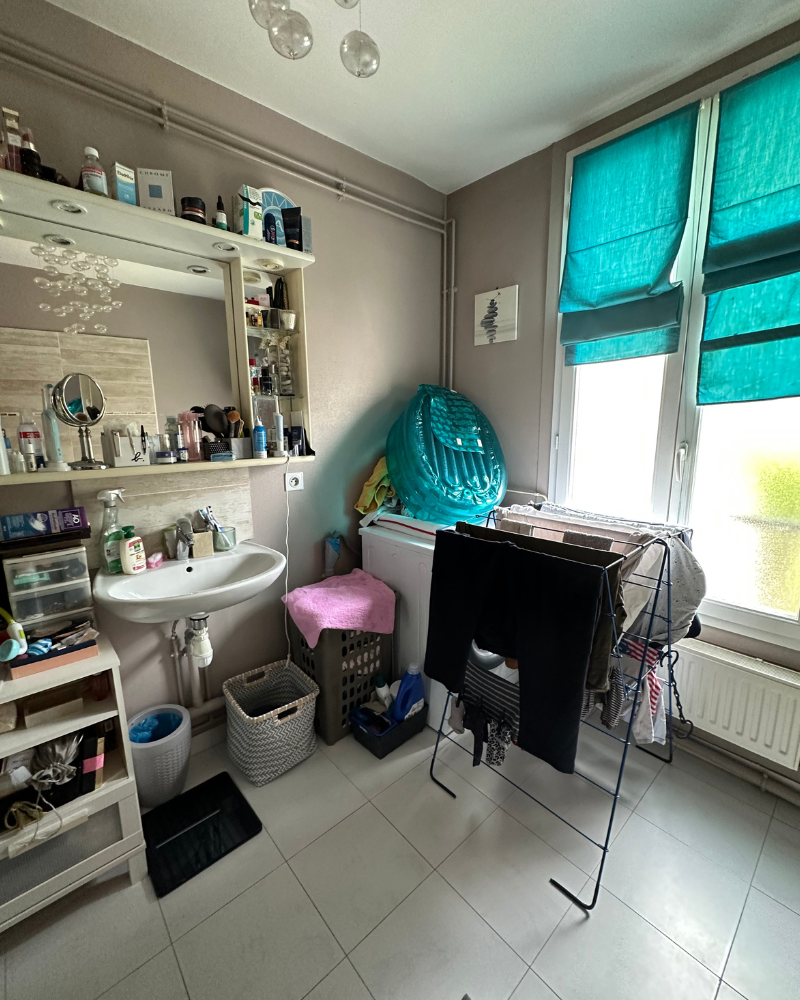 Une salle de bain encombrée avec des articles de toilette sur des étagères, un lavabo, un panier à linge, un séchoir avec des vêtements et un grand objet gonflable près d'une fenêtre avec des stores turquoise.