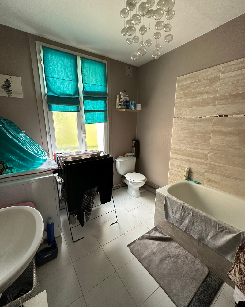 Une salle de bain avec baignoire, WC et lavabo. Un séchoir à linge est situé près de la fenêtre, qui est dotée de rideaux bleu sarcelle. Un plafonnier en forme de bulle est situé au-dessus. Les murs sont gris-beige.