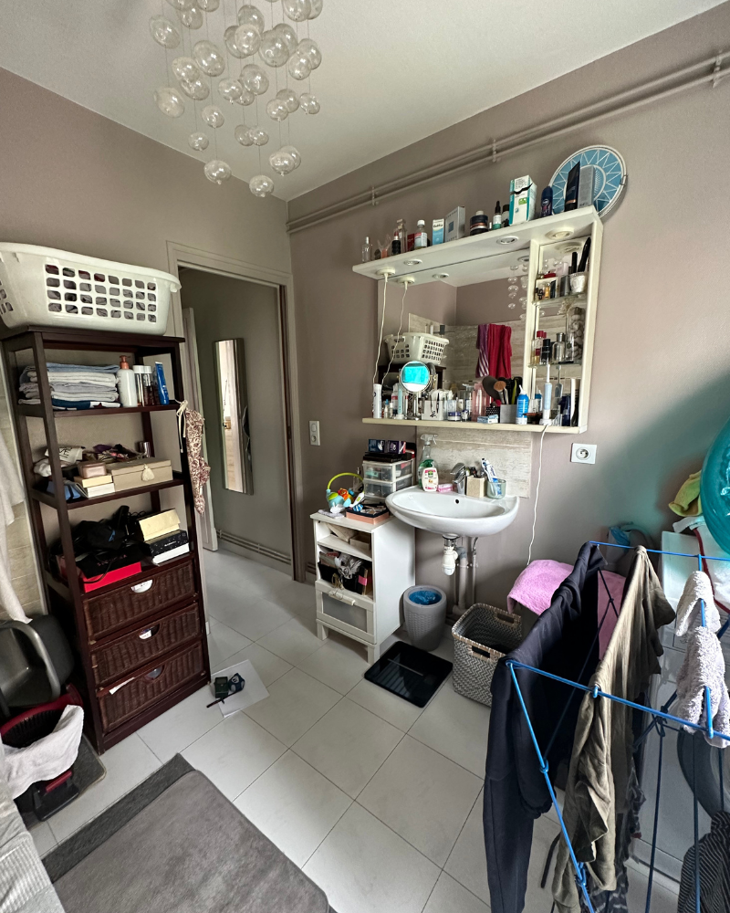 Une salle de bain avec un lavabo, une armoire à glace et des étagères remplies d'articles de toilette. Un séchoir à linge, un panier à linge et un luminaire suspendu sont également visibles.