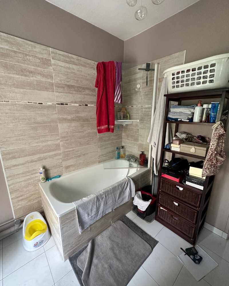 Une salle de bain avec une baignoire, des serviettes suspendues, une étagère avec des produits de toilette, un panier à linge au-dessus et un petit pot au sol.