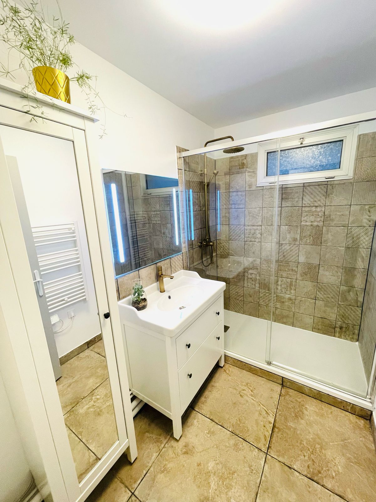Salle de bains avec douche vitrée, murs carrelés, meuble-lavabo blanc avec deux miroirs et armoire. Une plante en pot est posée sur l'armoire.