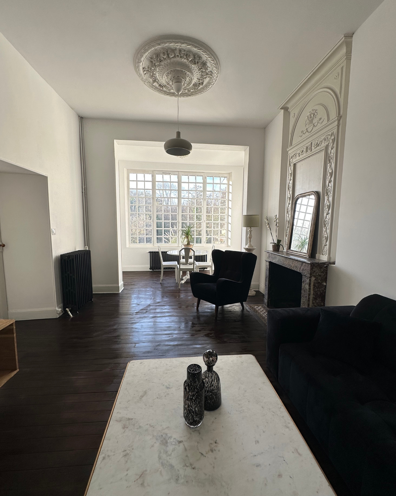 Salon spacieux avec parquet en bois foncé, cheminée en marbre, plafond décoratif et grandes fenêtres. Il comprend un canapé noir, un fauteuil et une petite table avec des chaises près de la fenêtre.