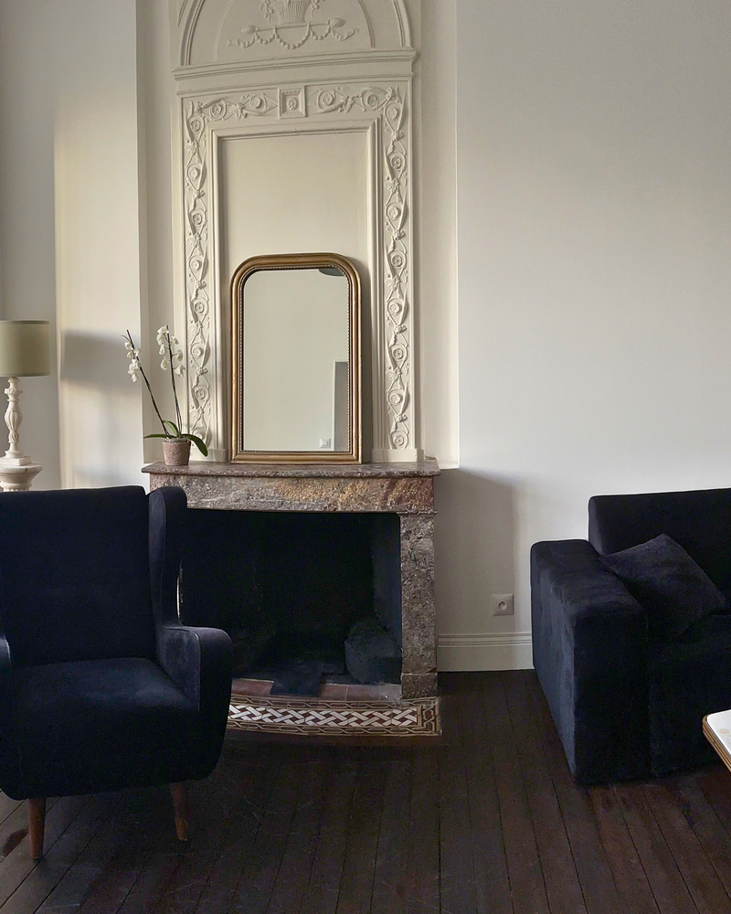 Élégant salon avec un grand miroir blanc orné au-dessus d'une cheminée en marbre, flanquée de deux fauteuils sombres. Une petite lampe et une plante sont posées sur le manteau. Parquet en bois foncé.