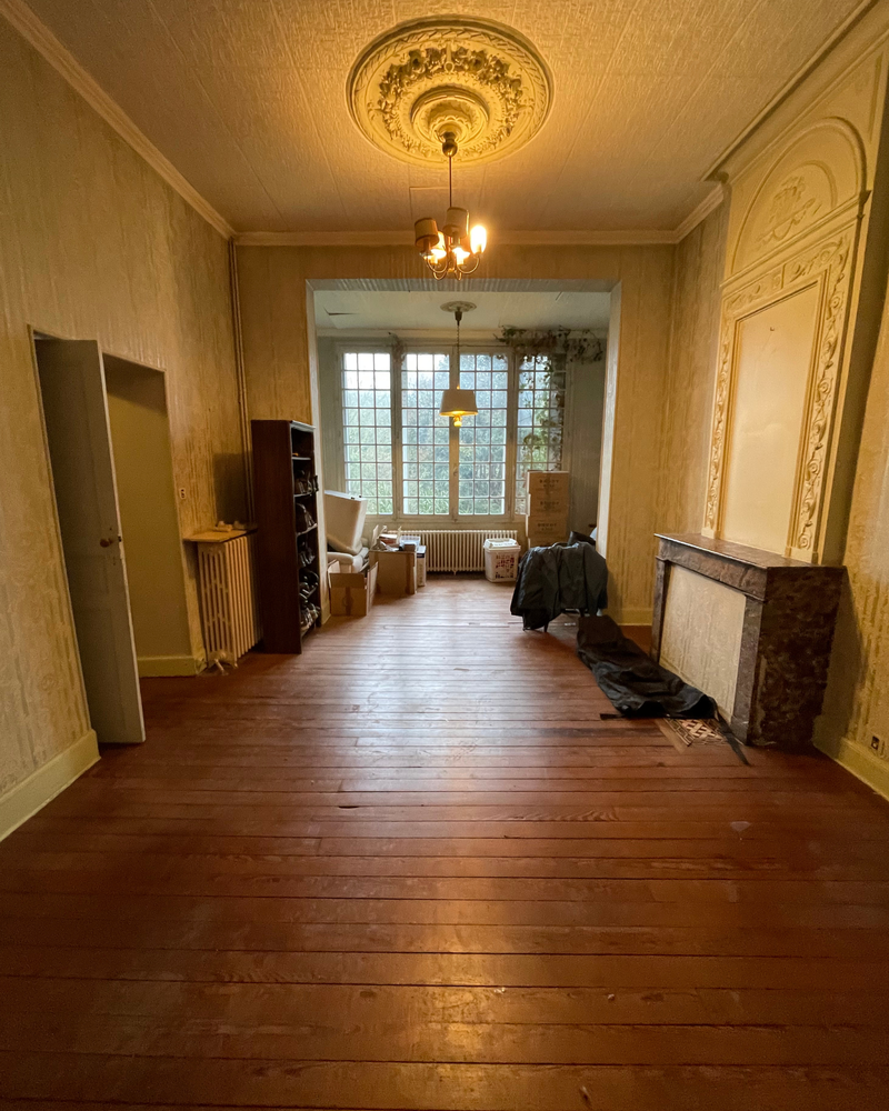 Salon_1_Avant_A Une pièce vintage avec parquet, plafond orné et cheminée. On y voit des boîtes, des étagères et un manteau, et de grandes fenêtres au fond laissent entrer la lumière naturelle.