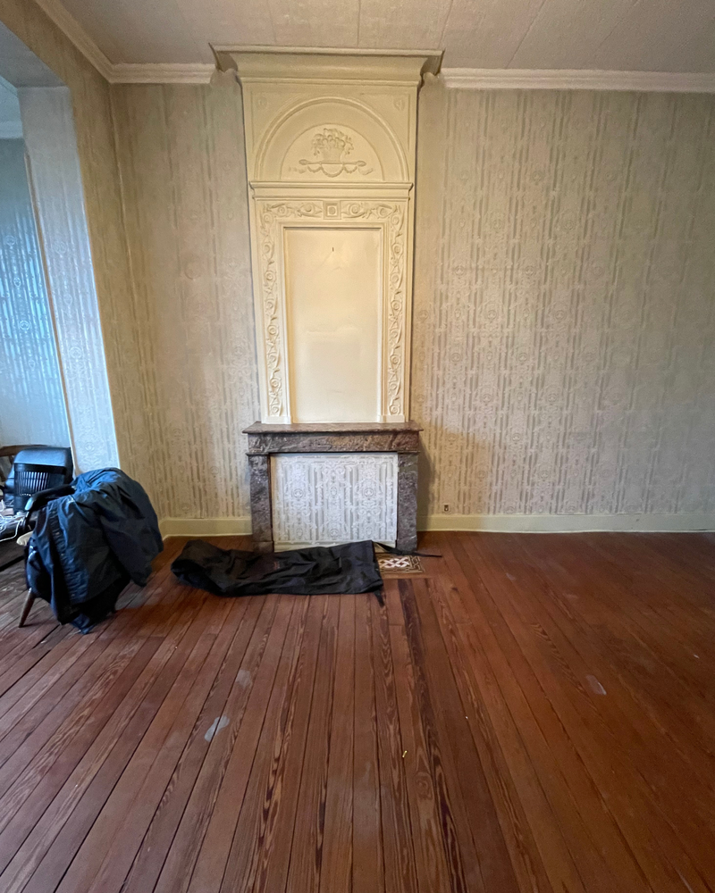 Salon_1_Avant_C Pièce vide avec parquet, boiseries décoratives, manteau de cheminée décoratif et tapis sombre au sol. Une veste noire est drapée sur une chaise dans un coin.