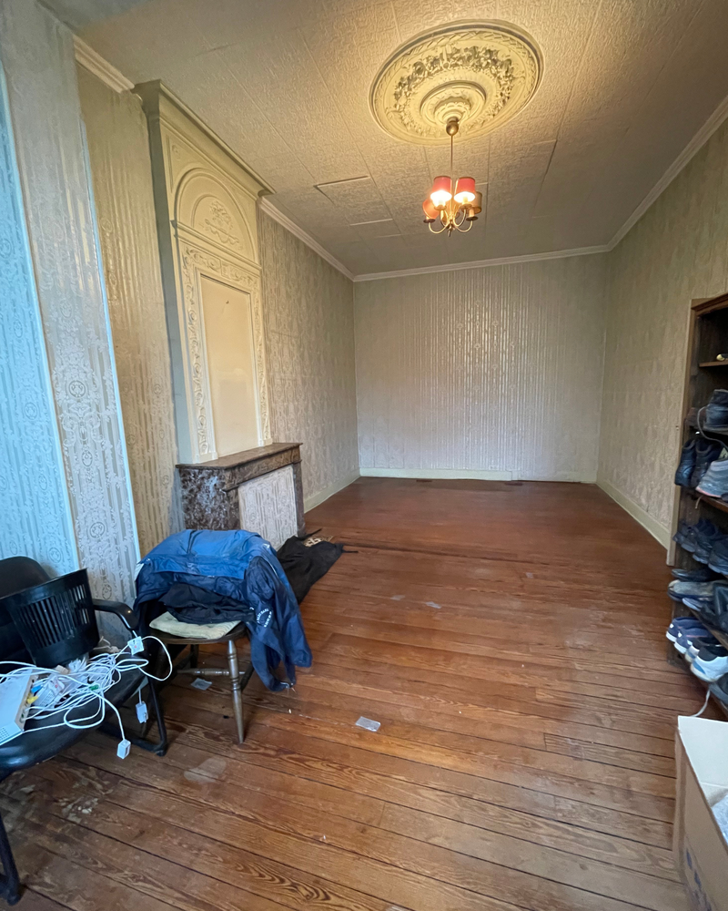 Salon_1_Avant_E Une pièce vide avec un parquet, un médaillon décoratif au plafond et un petit lustre. Un manteau de cheminée orne un mur. Une chaise avec des objets se trouve au premier plan à gauche.