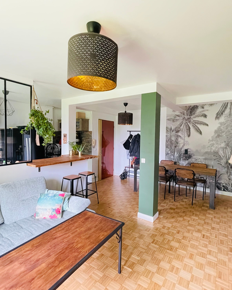 Espace de vie ouvert avec coin salon, table à manger et tabourets de bar. Murs aux accents verts, parquet, luminaires suspendus et fresque murale ornée de plantes tropicales ornent la pièce.