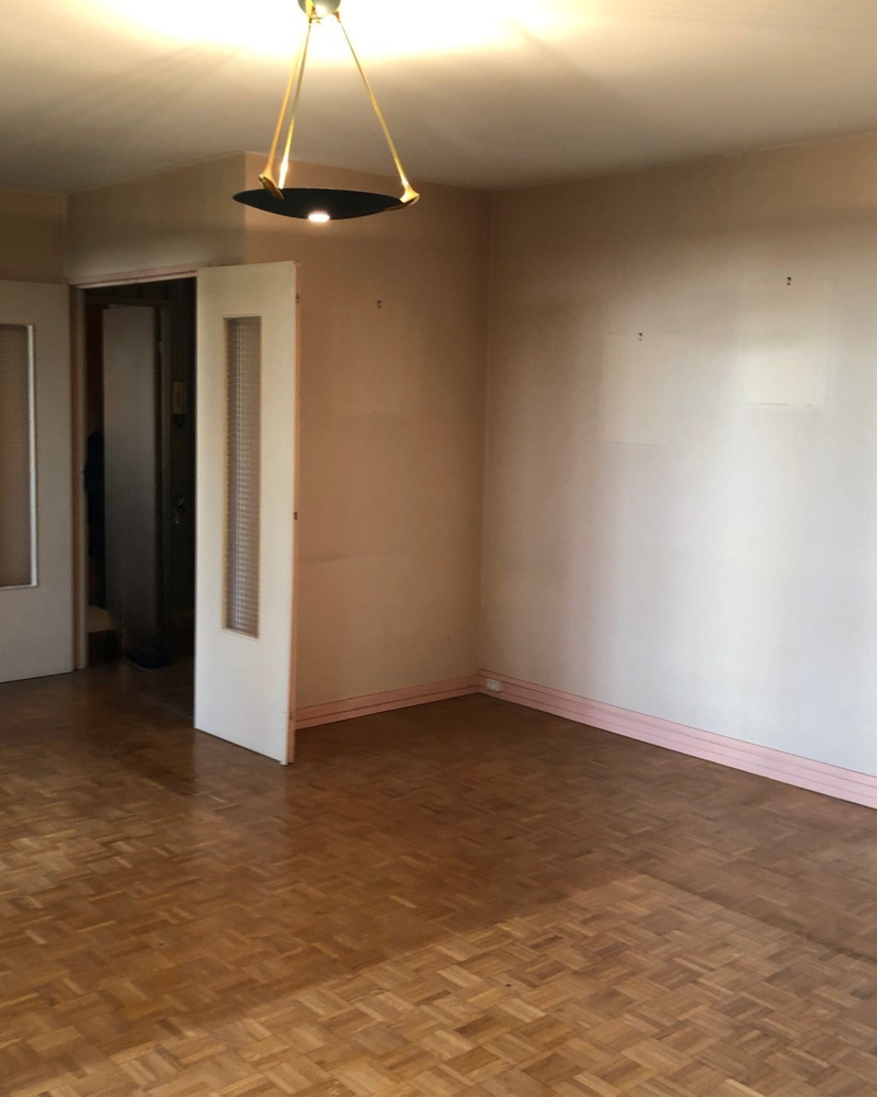 Salon_2_Avant_A Une pièce vide aux murs beiges, au parquet et au plafonnier moderne. Une porte entrouverte à gauche révèle un petit couloir.