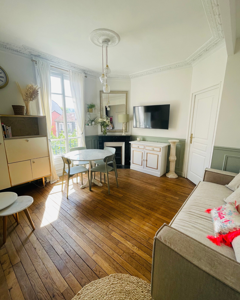 Un salon lumineux avec une petite table à manger, une télévision murale, un canapé et du parquet. Une grande fenêtre avec rideaux laisse entrer la lumière naturelle. D'élégantes moulures au plafond et une horloge décorative sont présentes.