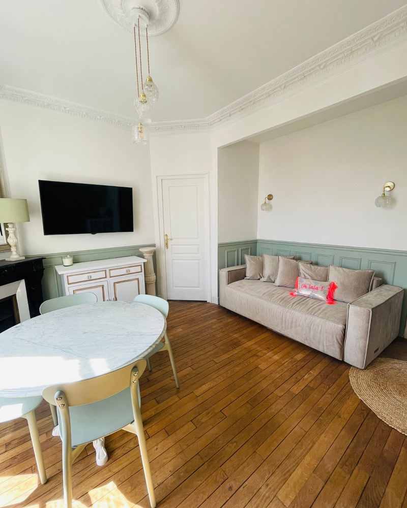 Salon lumineux avec canapé, télévision murale, table ronde et chaises sur parquet. Murs blancs avec lambris verts et moulures décoratives au plafond.