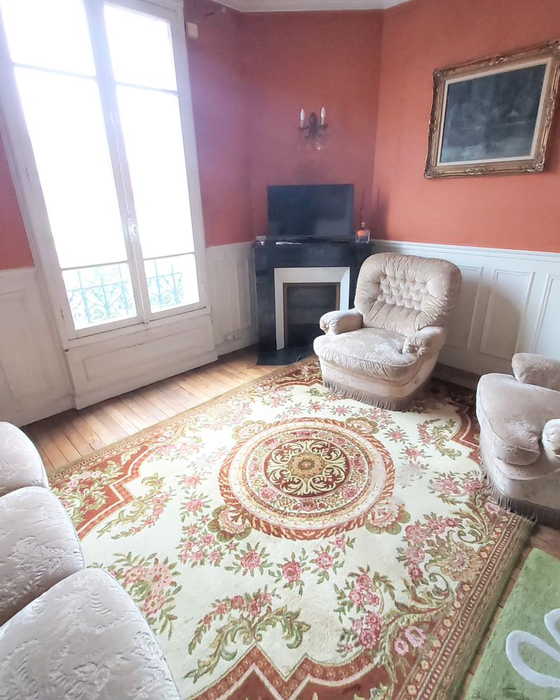 Salon_3_Avant_A Un salon avec des murs orange, un grand tapis fleuri, des chaises rembourrées, une cheminée et une télévision près d'une fenêtre avec des rideaux.