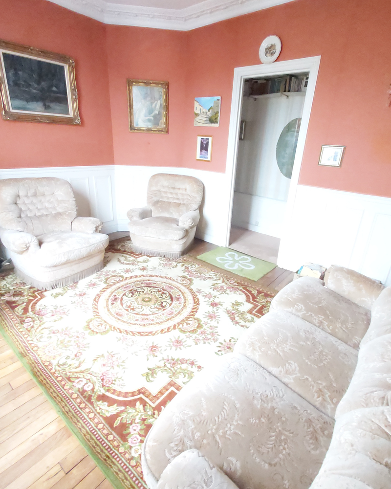 Salon_3_Avant_C Un salon de style vintage avec du papier peint à fleurs, un tapis orné, trois fauteuils beiges et un canapé assorti. Des tableaux et une photo sont accrochés aux murs. Une porte mène à une autre pièce.
