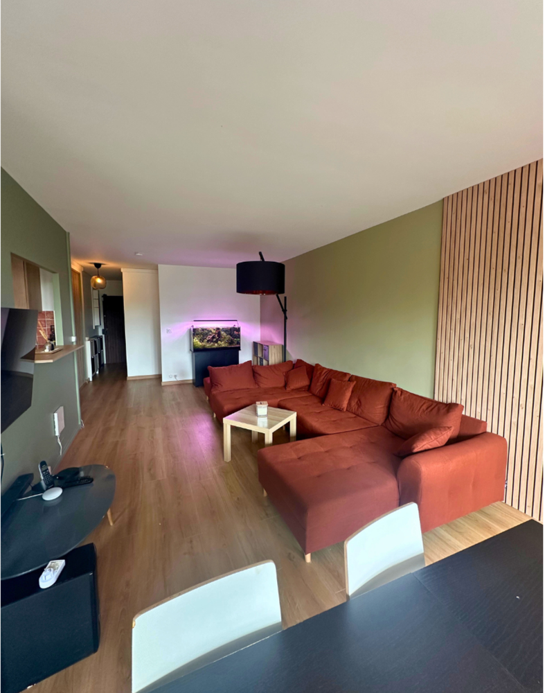 Un salon moderne avec un grand canapé d'angle marron, du parquet et une télévision murale. La pièce est agrémentée d'un mélange de lumière tamisée et de boiseries.