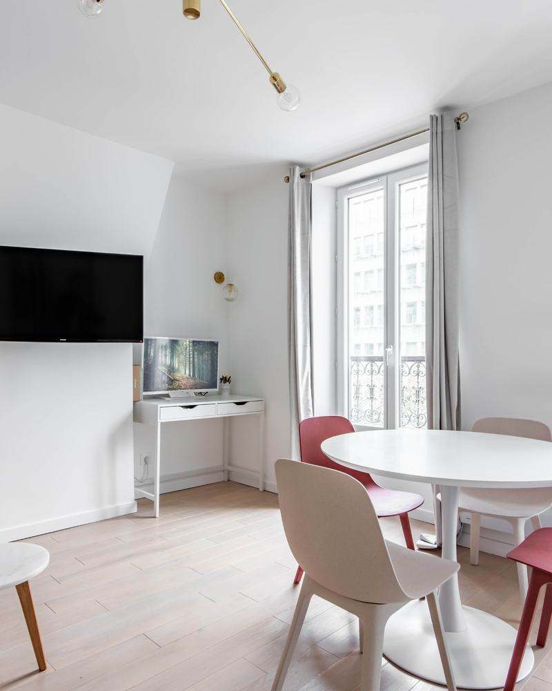 Chambre moderne avec une table ronde blanche, quatre chaises roses et blanches, une télévision murale, un petit bureau avec un écran, un parquet en bois clair et une grande fenêtre avec des rideaux gris.