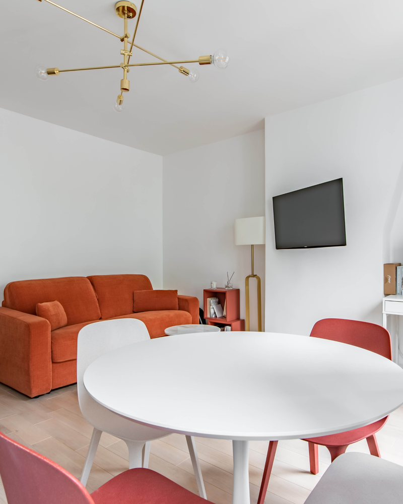 Salon moderne avec un canapé rouge, une table à manger ronde blanche, des chaises rouges et blanches, une télévision murale, un lustre doré et un petit lampadaire.