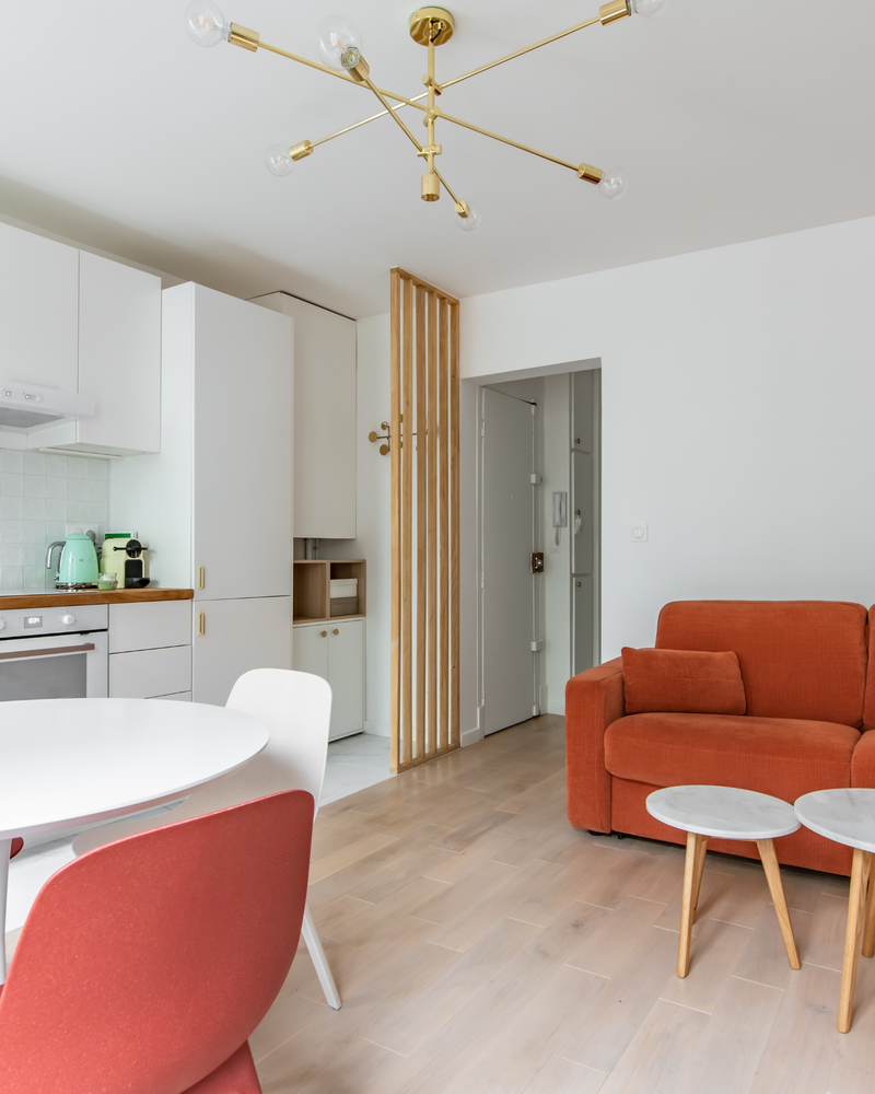 Espace de vie moderne avec armoires blanches, canapé rouge, table à manger ronde blanche avec chaises rouges et blanches, deux petites tables rondes et un lustre doré. Parquet clair.