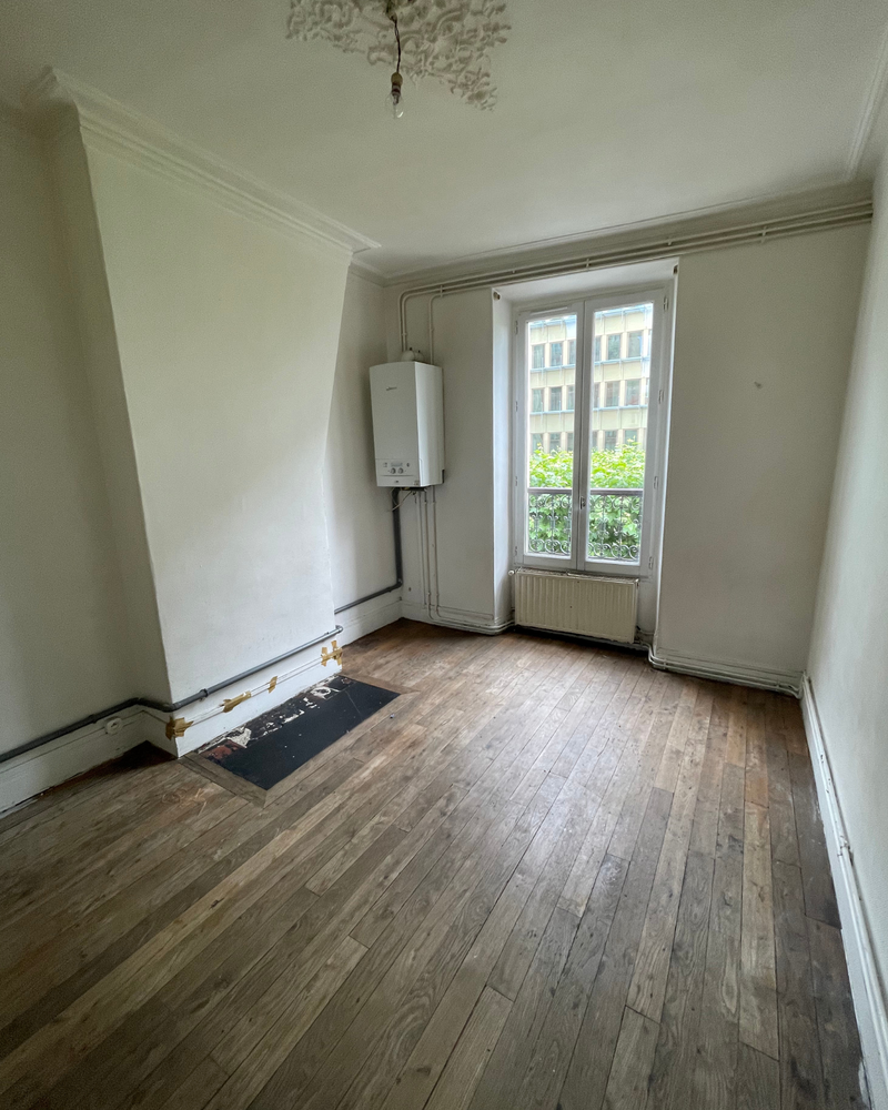 Salon_5_Avant_A Pièce vide avec parquet, murs blancs et fenêtre donnant sur un bâtiment. Une chaudière est fixée sur un mur. La pièce présente des canalisations apparentes et un simple plafonnier.