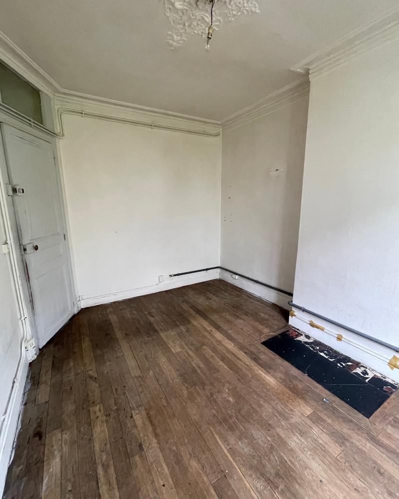 Salon_5_Avant_C Une pièce vide avec des murs blancs, du parquet, un petit morceau de sol manquant et une porte blanche sur la gauche.