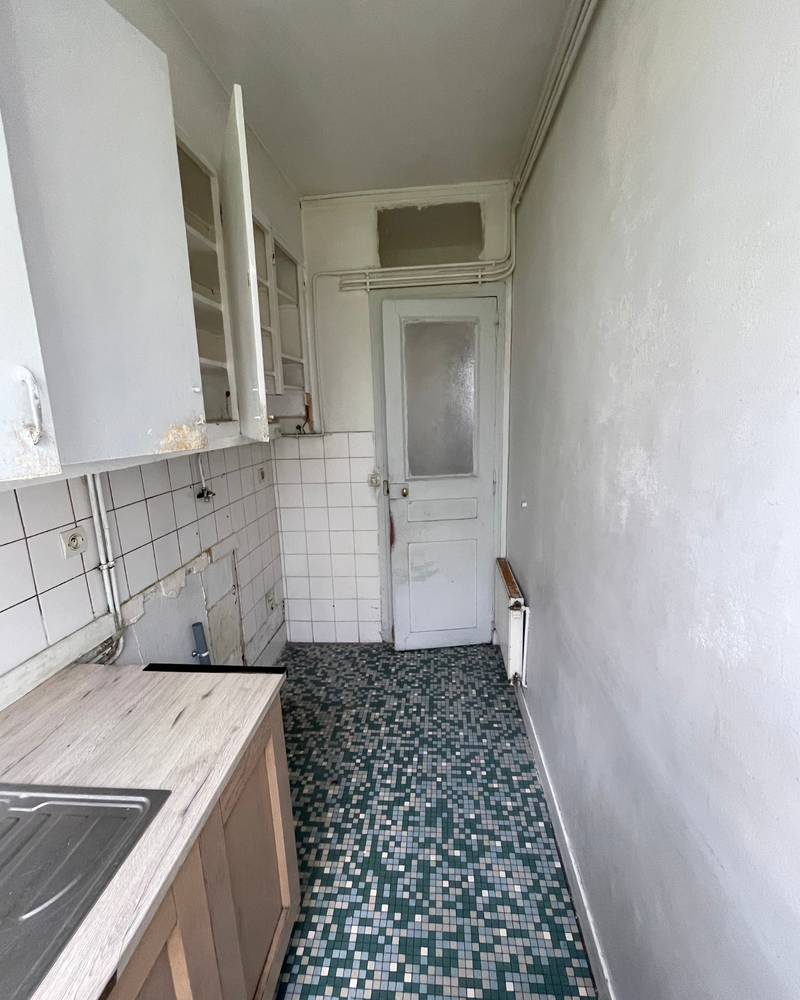 Salon_5_Avant_E Cuisine étroite et sombre avec sol carrelé, placards blancs et murs délabrés. Un placard ouvert révèle des étagères vides. Les éviers et les plans de travail semblent usés. La porte est au fond.