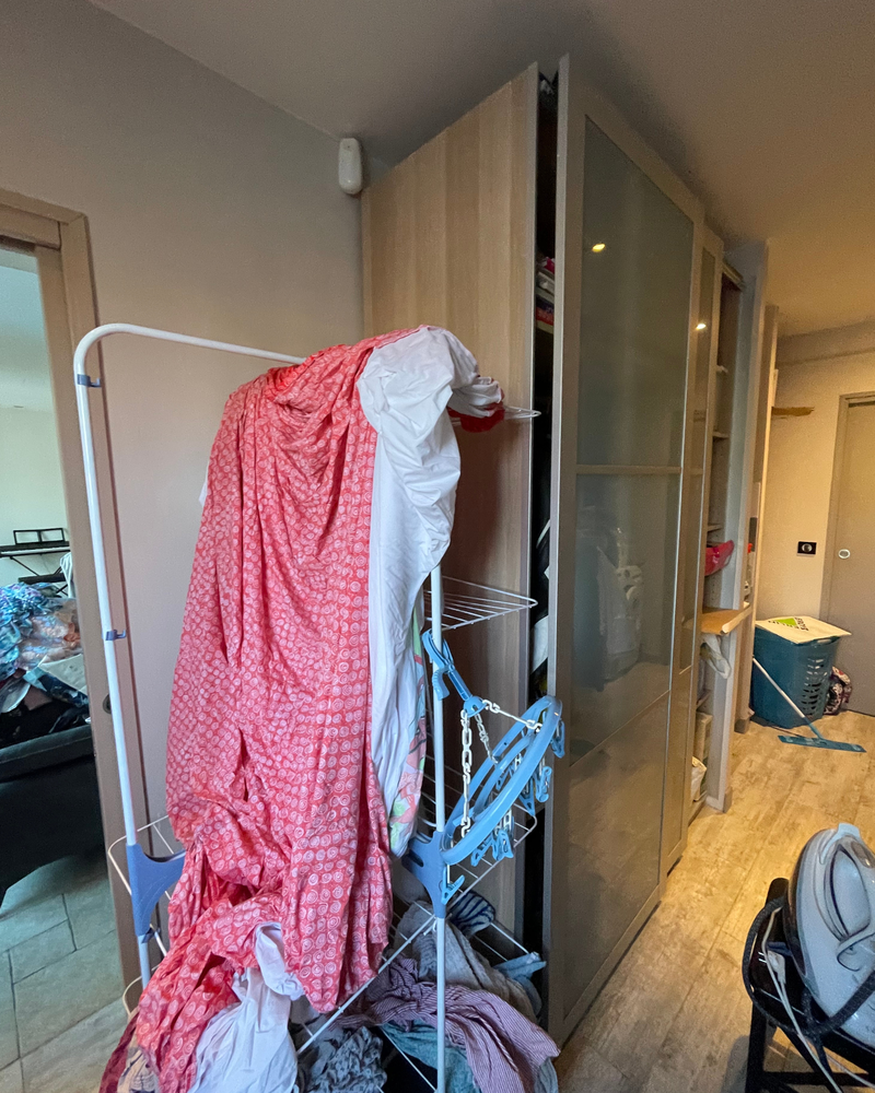 Un séchoir recouvert d'un tissu rouge et blanc se trouve à côté d'une armoire dans une pièce encombrée où l'on voit du linge et divers articles.