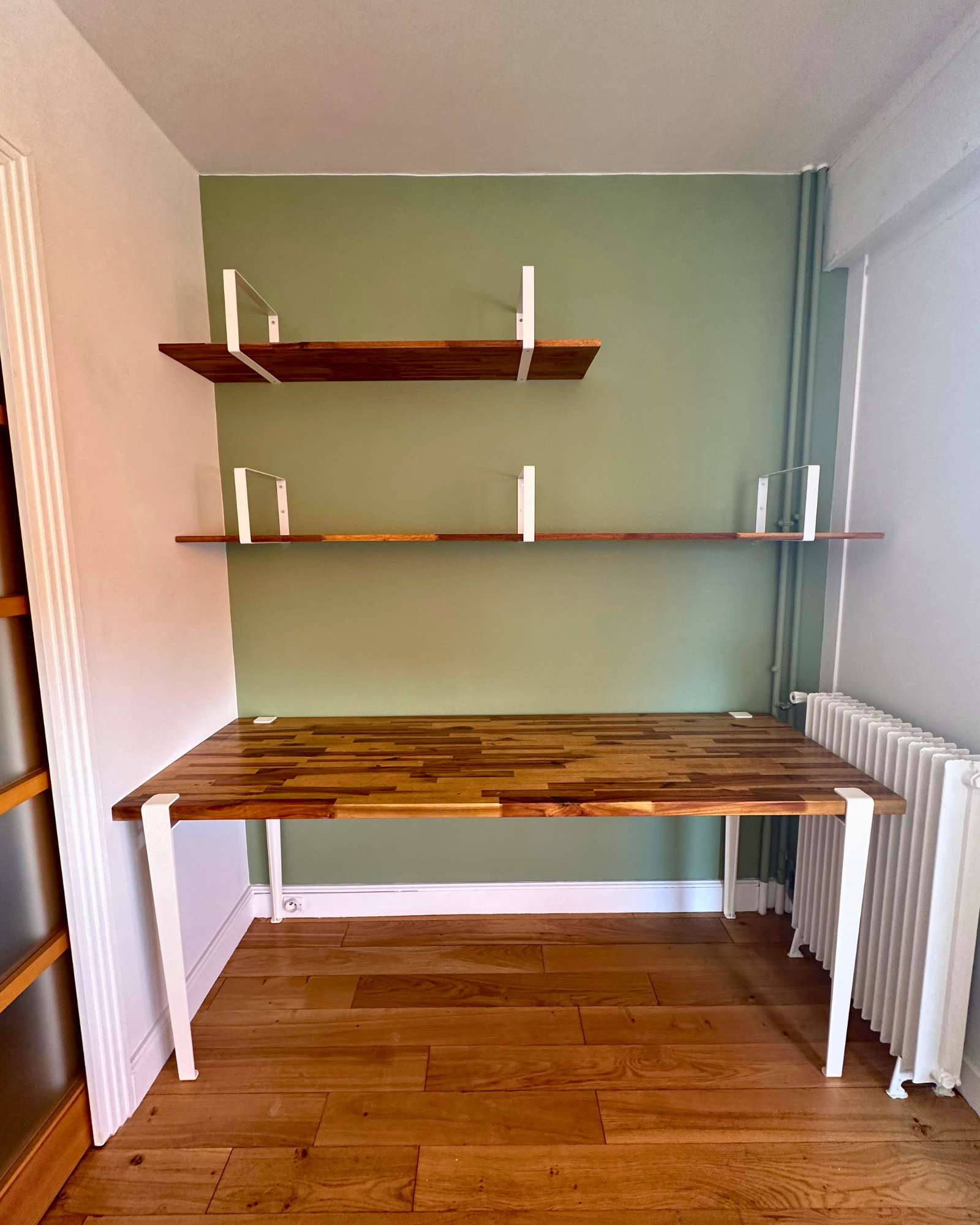 Un bureau en bois aux pieds en métal blanc est placé contre un mur végétalisé, surmonté de deux étagères en bois. Un radiateur blanc trône sur le côté droit.