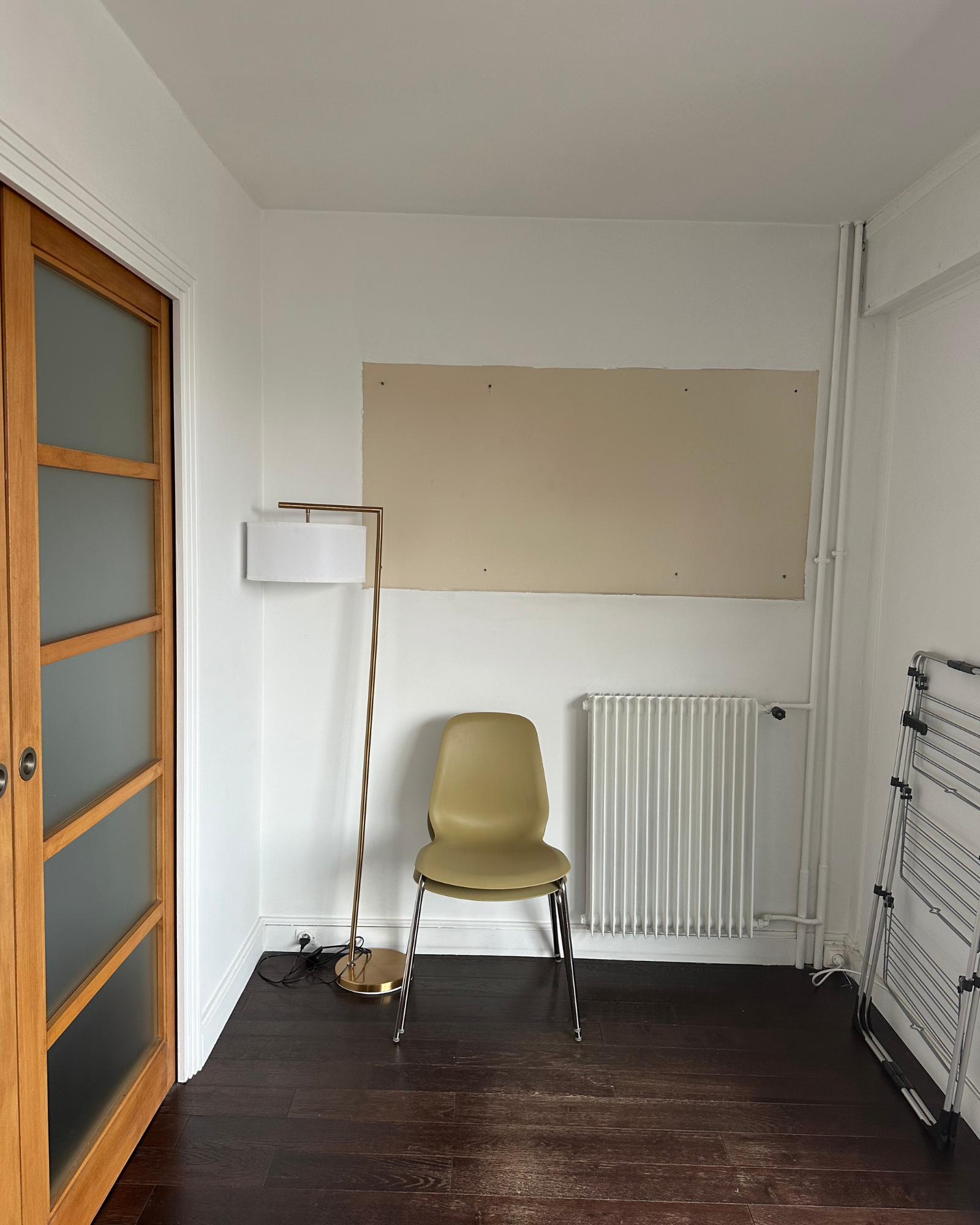 Une chaise beige, un lampadaire et un radiateur sont disposés contre un mur blanc avec une zone rectangulaire vide. On y voit également un séchoir pliable et une porte coulissante en bois.