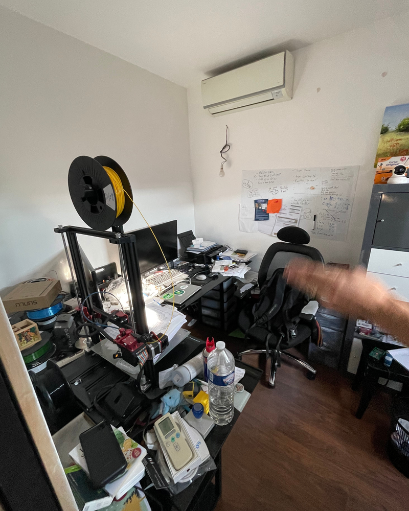 Bureau_2_Avant_C Un bureau à domicile encombré avec une imprimante 3D, un ordinateur, divers appareils électroniques, des papiers et une chaise de bureau. Un tableau blanc avec des notes et un climatiseur au mur.