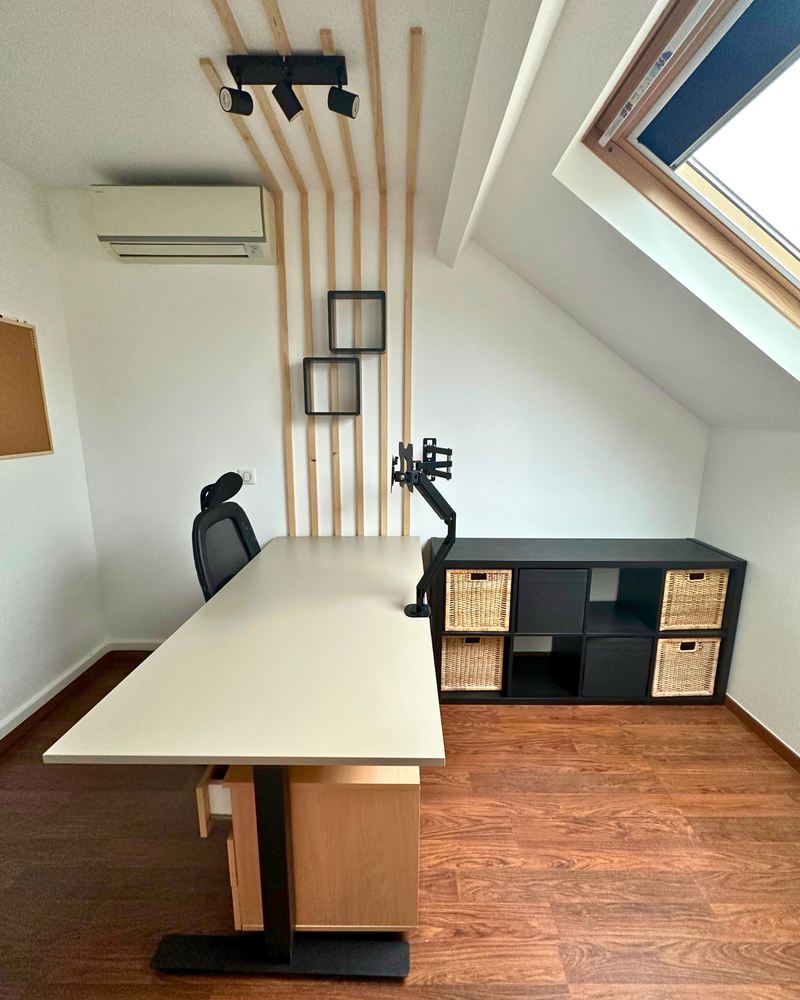Bureau à domicile minimaliste avec un bureau blanc, une chaise noire, un bras de moniteur mural, des étagères avec des paniers en osier et des accents muraux en bois sous une lucarne.