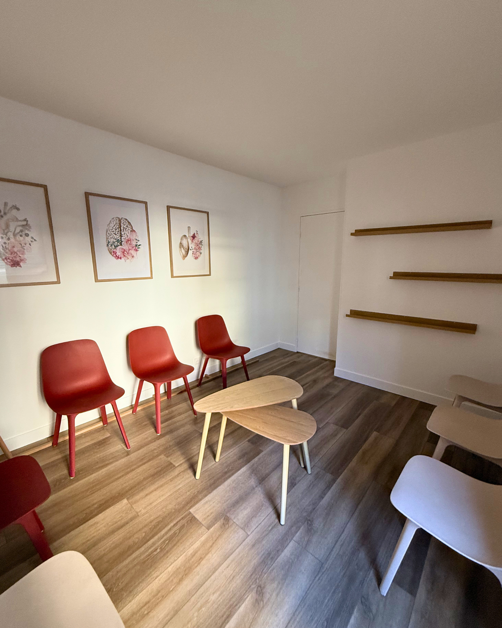 Une petite salle d'attente avec des chaises rouges et beiges, des tables en bois, des œuvres d'art encadrées au mur et des étagères murales vides.