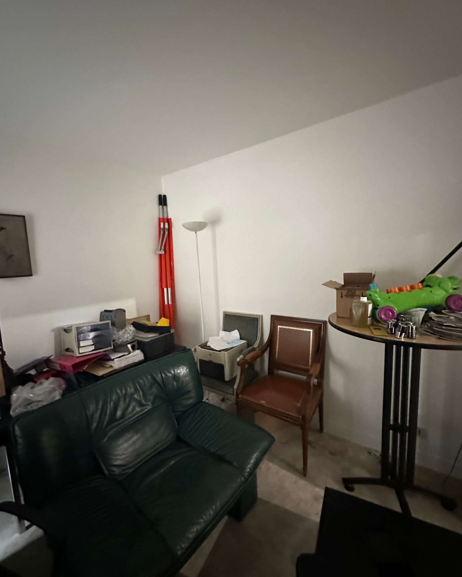 Une pièce encombrée avec un canapé vert, une chaise en bois, de hauts poteaux rouges, un lampadaire et des boîtes sur une table ronde. Un assortiment d'objets divers est éparpillé un peu partout.