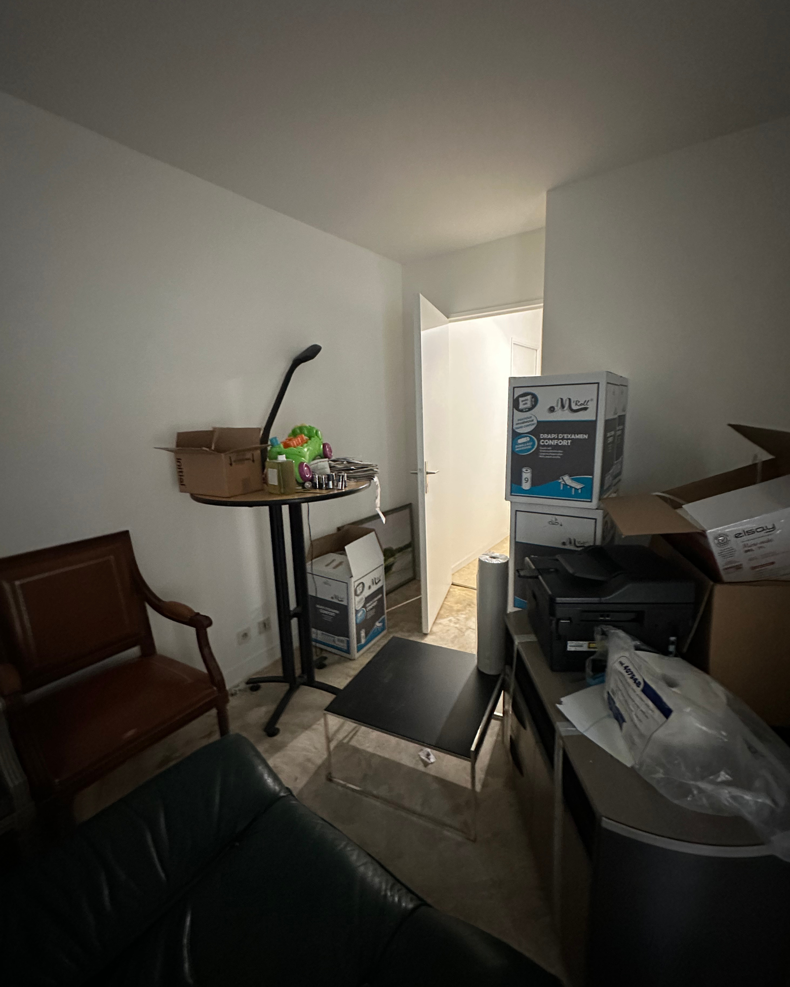 Une pièce encombrée avec des cartons, une chaise en bois, une table ronde avec des objets et une porte ouverte menant à un couloir éclairé.