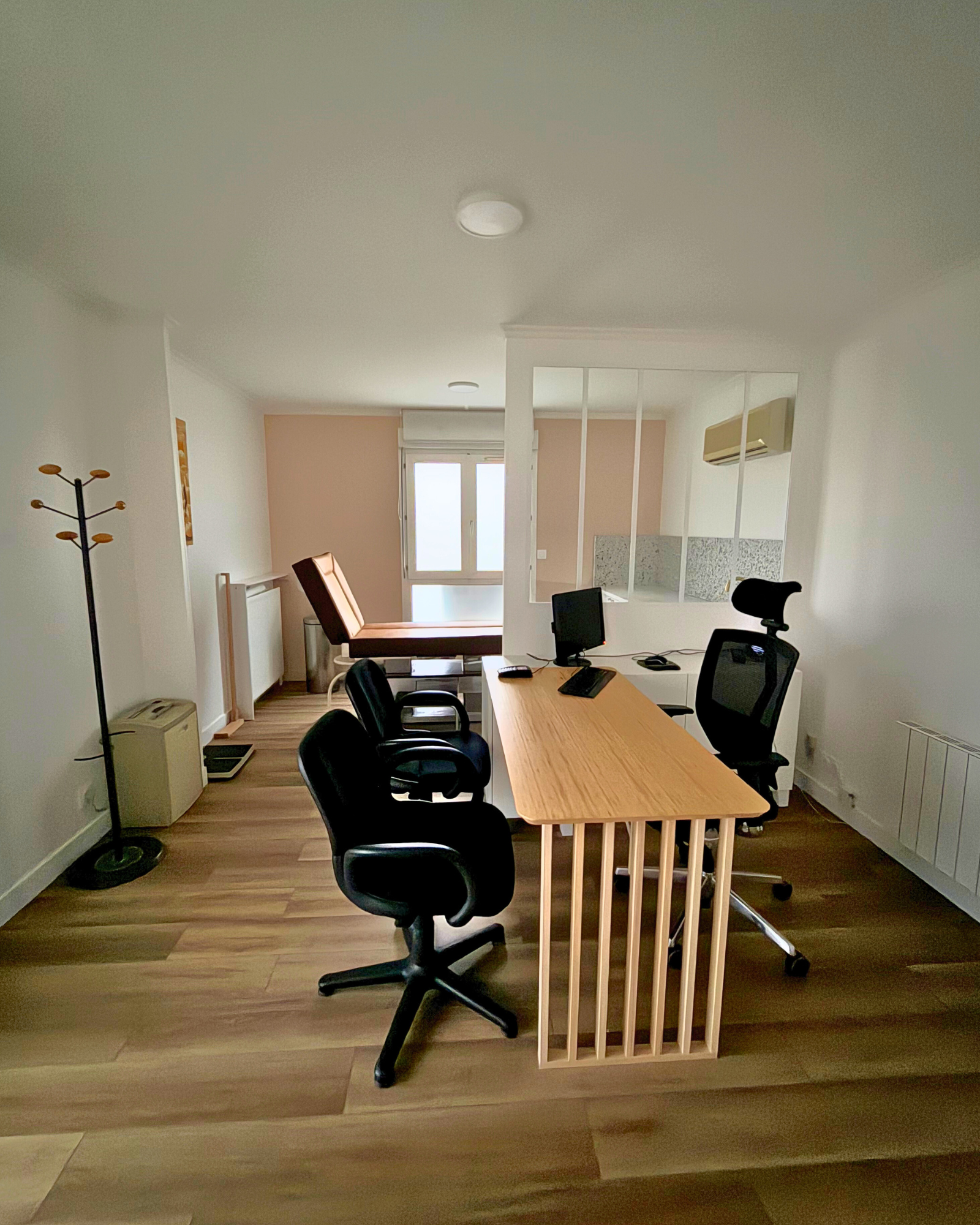 Espace de travail avec un bureau en bois, deux chaises noires, un écran d'ordinateur et un clavier. La pièce est dotée de parquet, d'un porte-manteau et de grandes fenêtres en arrière-plan.