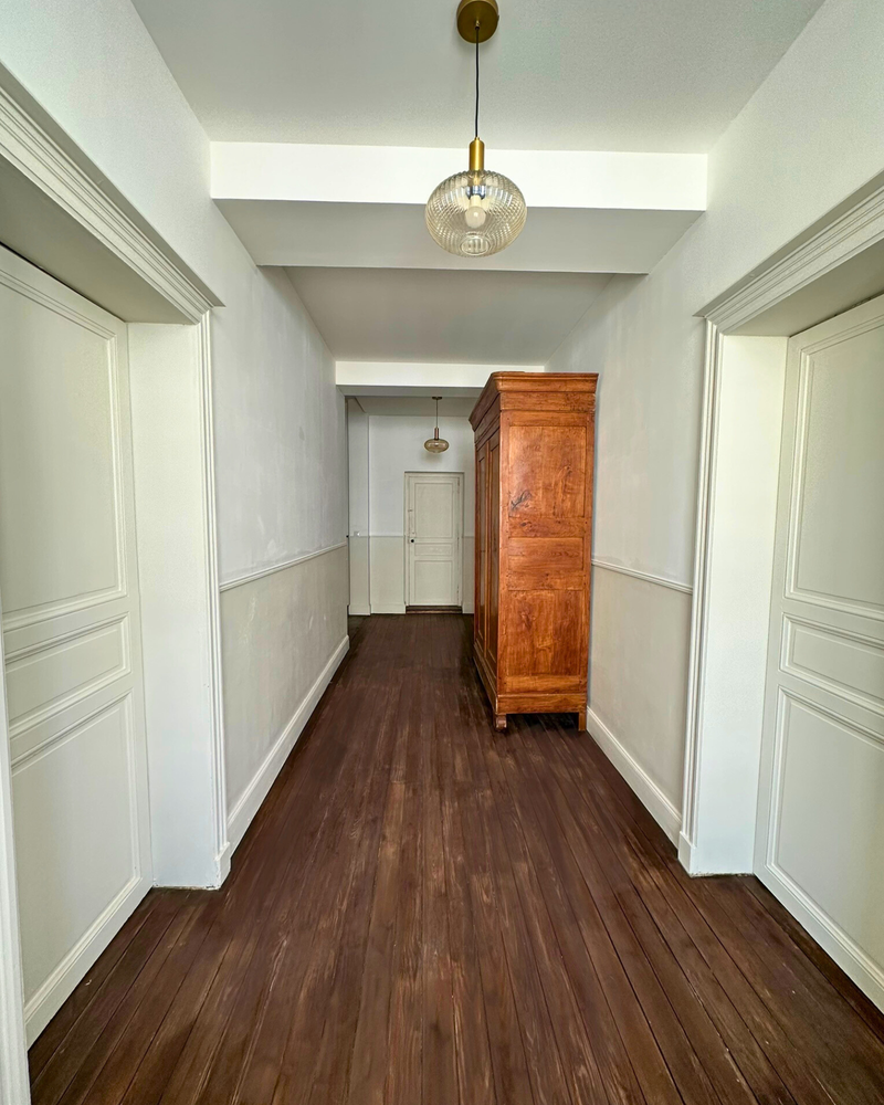 Un couloir étroit avec parquet, murs blancs, deux luminaires suspendus et une grande armoire en bois près du centre. Plusieurs portes fermées bordent le couloir.