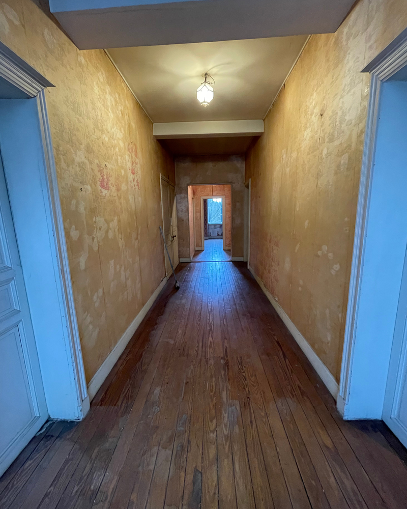Couloir_1_Avant_A Un long couloir étroit avec des planchers en bois usés et des murs délavés et irréguliers, éclairé par une seule ampoule au plafond, mène à des pièces avec des portes ouvertes et une fenêtre au fond.