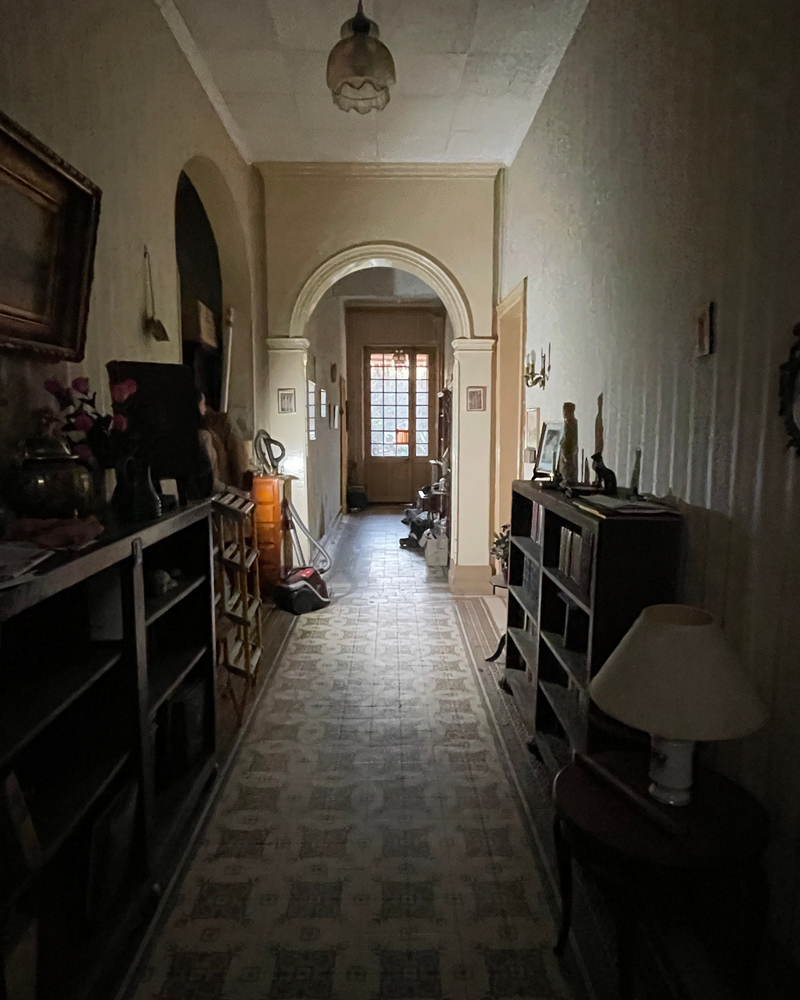 Couloir_2_Avant_A Un couloir faiblement éclairé avec du carrelage à motifs, des étagères et des meubles de chaque côté, menant à une porte avec des vitres au fond.