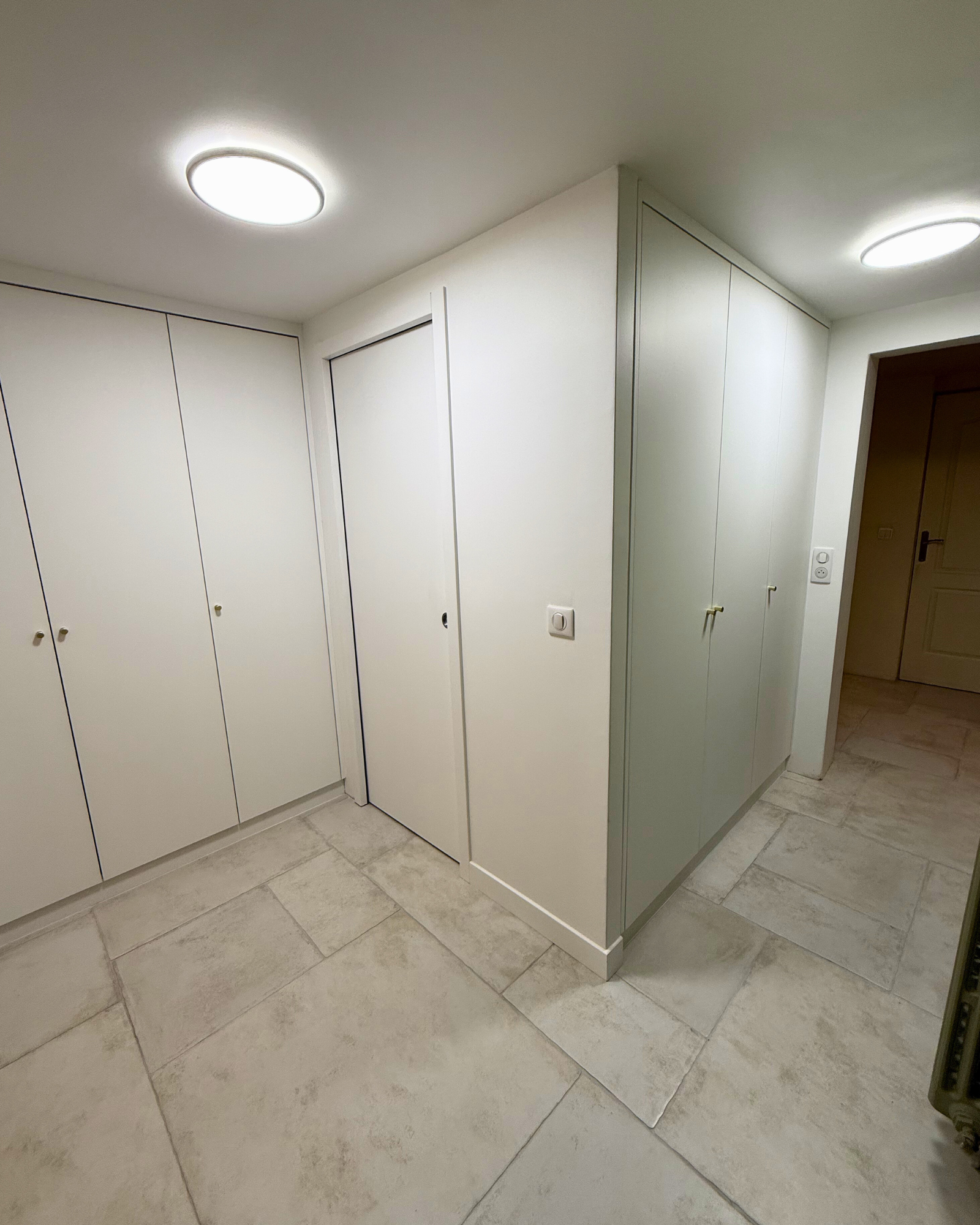 Un couloir avec un carrelage de couleur claire, des murs blancs, des placards intégrés avec des portes fermées et deux plafonniers ronds.