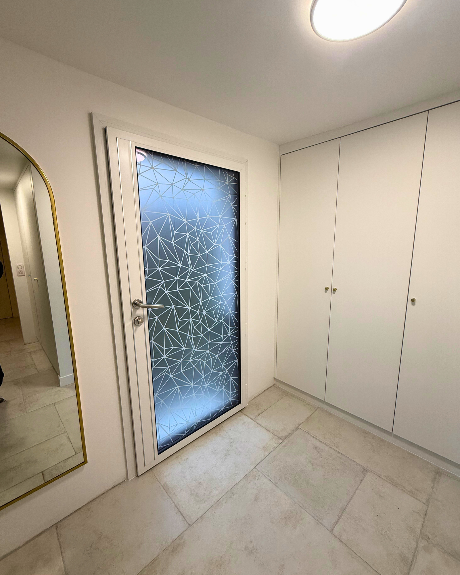 Un couloir avec une porte en verre dépoli présentant un motif géométrique, des armoires blanches, un grand miroir mural et un sol carrelé.