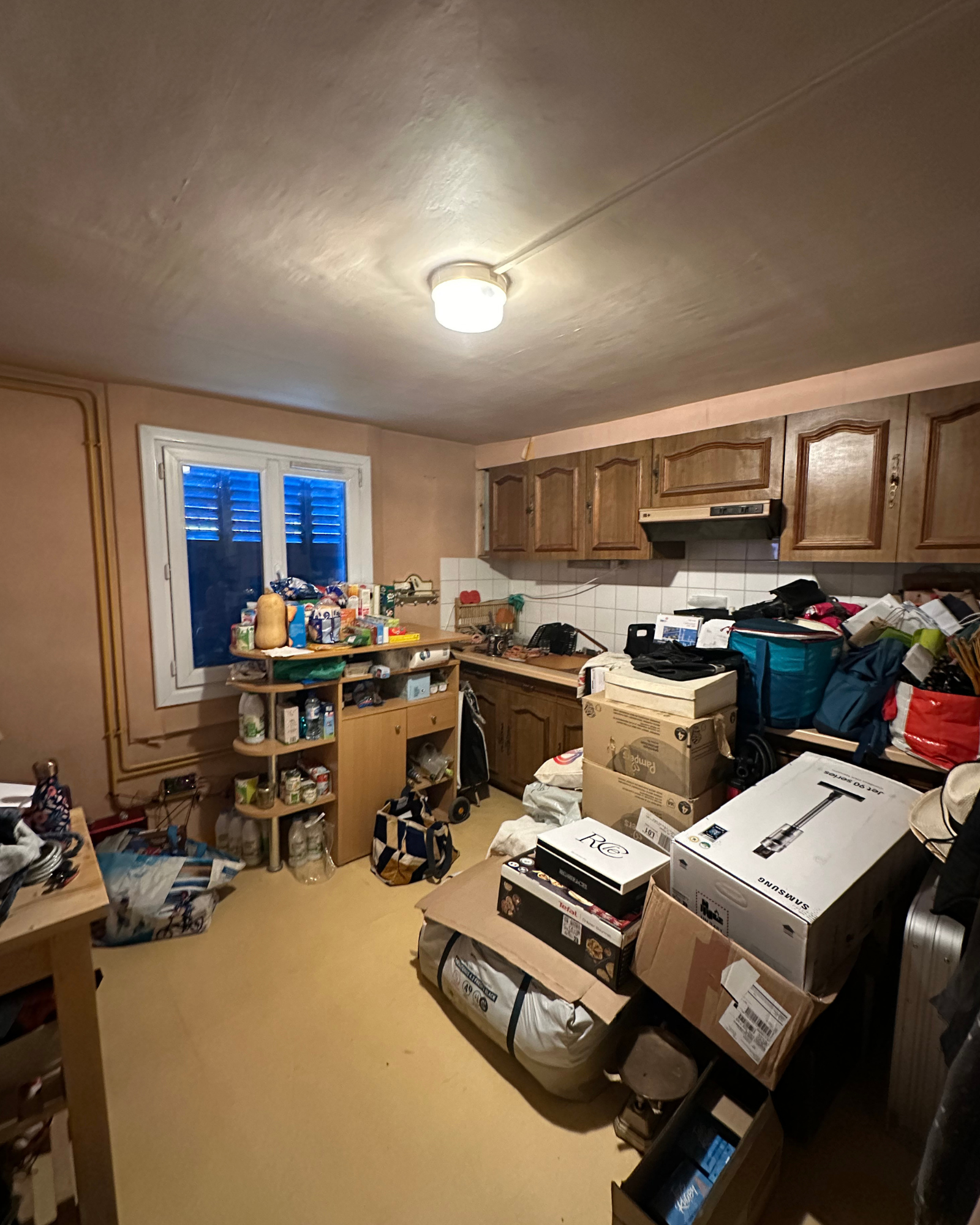 Couloir_3_Avant_A Une cuisine encombrée avec des armoires en bois, des boîtes en carton, des sacs et des articles ménagers éparpillés sur le sol et les comptoirs sous un plafonnier.