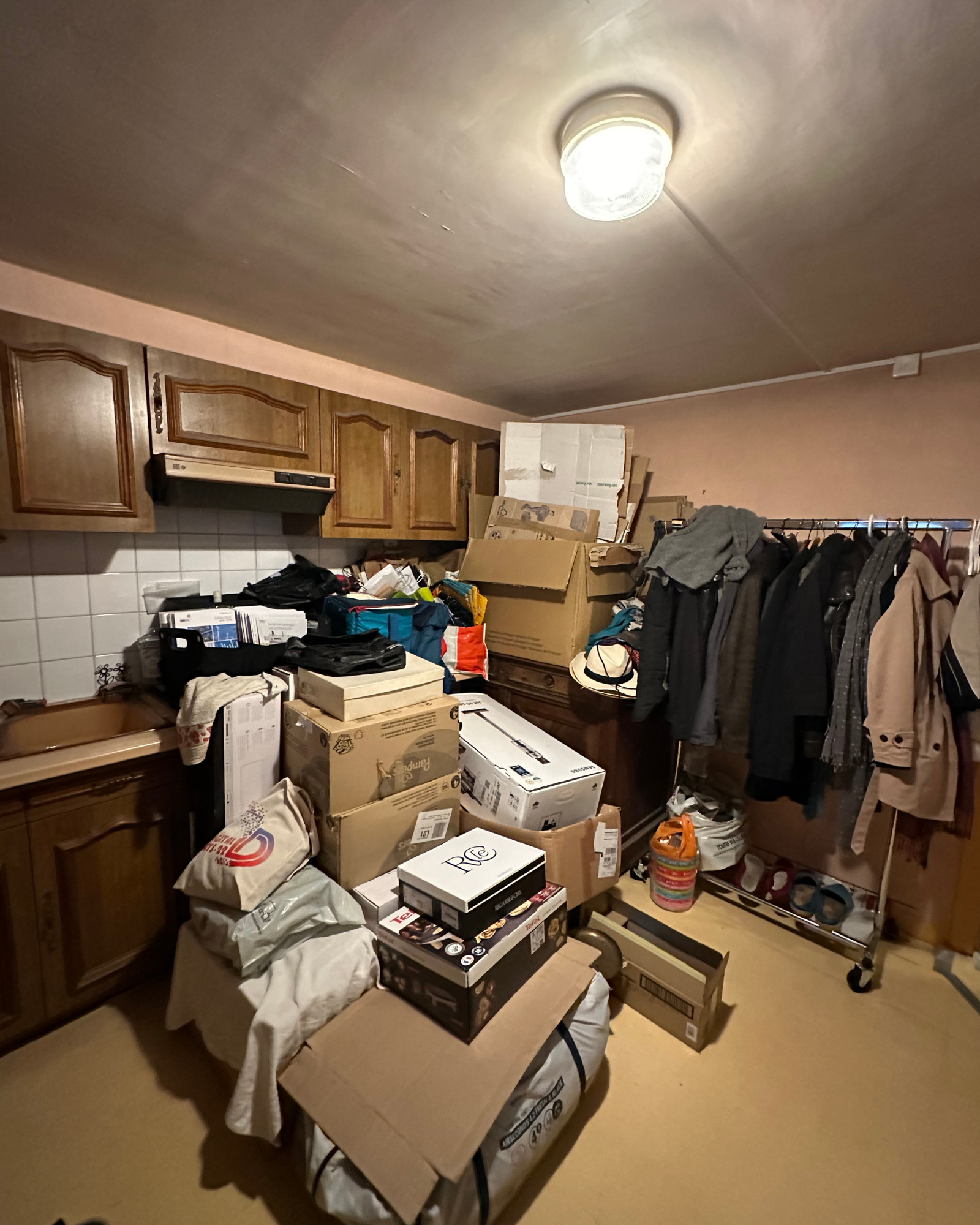 Couloir_3_Avant_C Une pièce encombrée de cartons, de vêtements sur un portant, de chaussures et de divers articles ménagers empilés sur le sol et le comptoir sous des armoires en bois.
