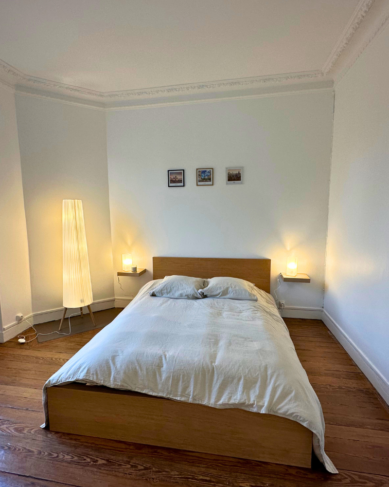 Un lit soigneusement fait avec une literie beige est centré dans une pièce avec du parquet, deux petites étagères murales avec des lampes, un grand lampadaire et trois tableaux encadrés au-dessus de la tête de lit.