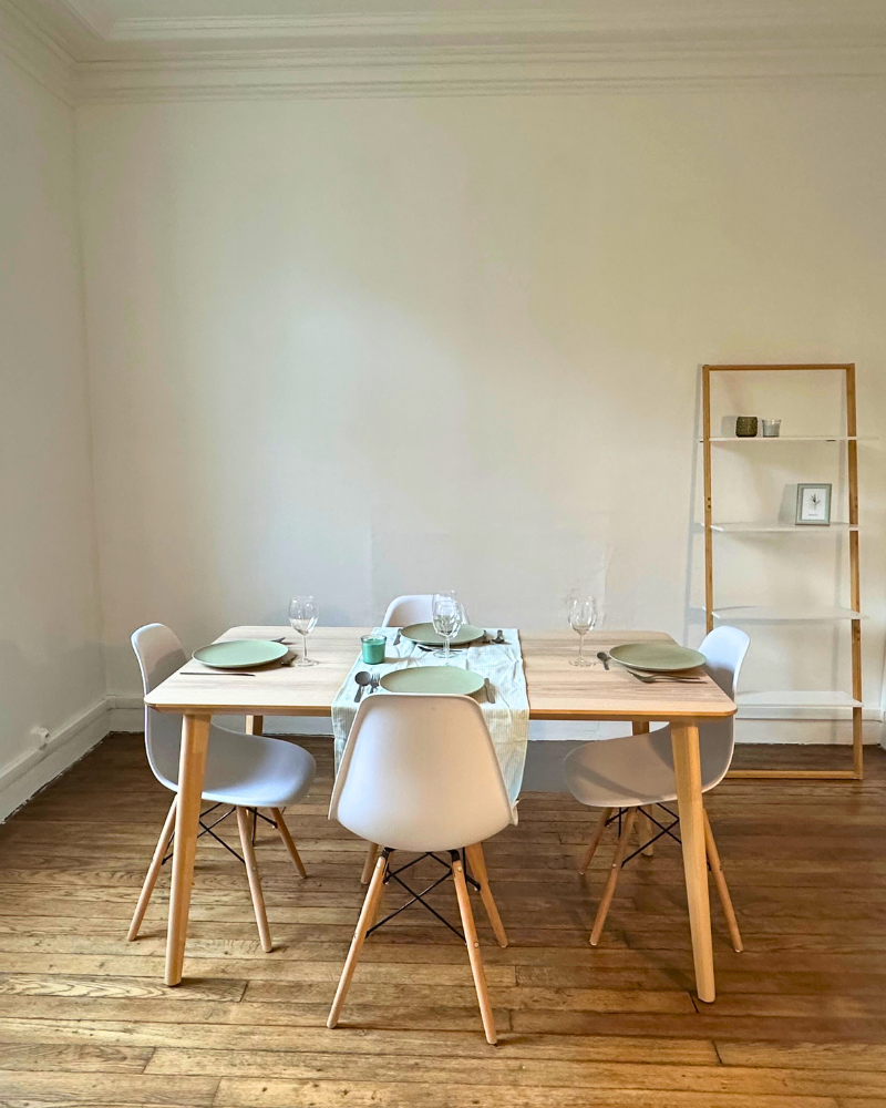 Une petite table à manger en bois pour quatre personnes avec des chaises de couleur claire, de la vaisselle simple et une étagère minimaliste contre un mur uni sur un plancher en bois.