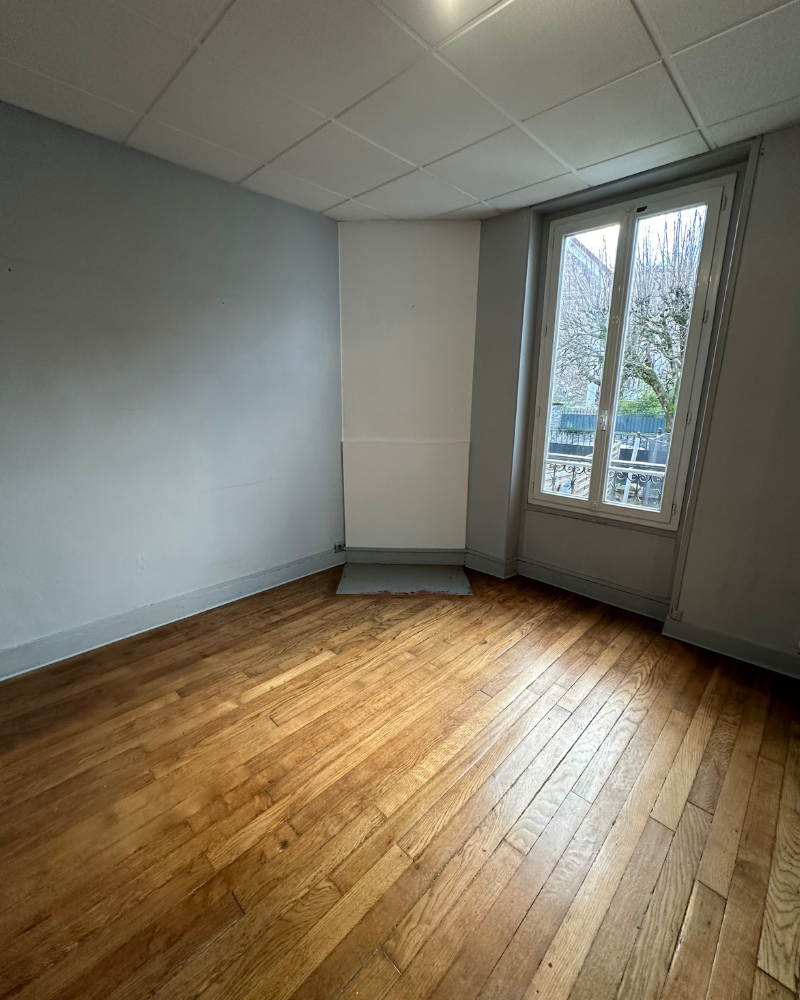 HS_1_Avant_A Pièce vide avec des murs gris clair, un parquet, des carreaux de plafond suspendu et une fenêtre montrant des arbres et une rue à l'extérieur.